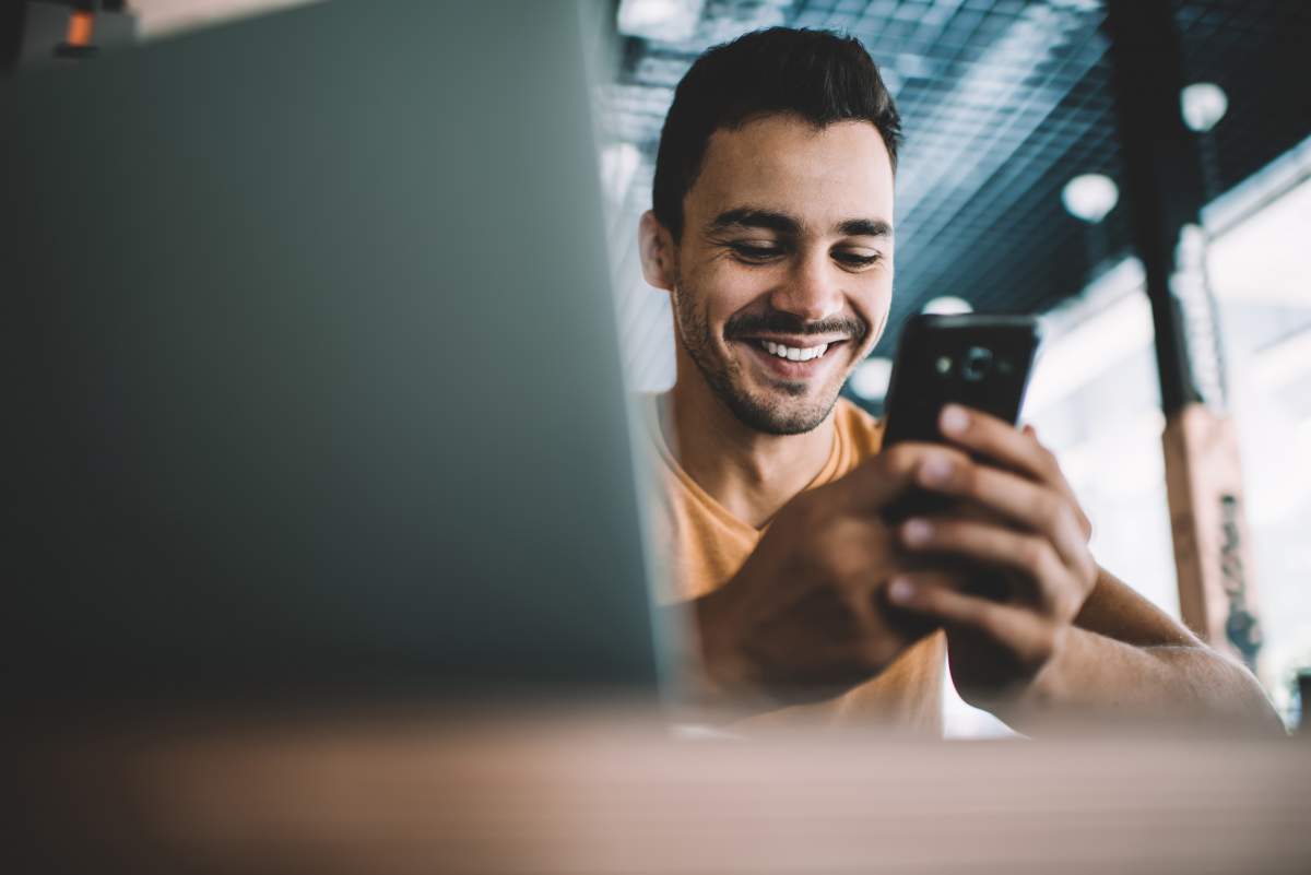 Junger Mann blickt auf ein Smartphone und lächelt, im verschwommenen Vordergrund ein Laptop-Screen
